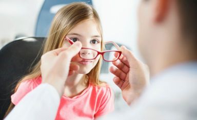 Little brow eyed girl visiting optician.She's having appointment for testing new glasses.Middle aged male optician is gently adjusting new frame and glasses on girl's face.