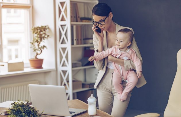 It is not easy to be a working mom! Young beautiful businesswoman talking on mobile phone and looking at laptop while standing with her baby girl at her working place