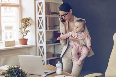 It is not easy to be a working mom! Young beautiful businesswoman talking on mobile phone and looking at laptop while standing with her baby girl at her working place