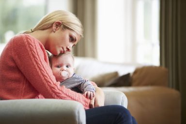 mother in pink sweater sitting on couch with a baby with pacifier