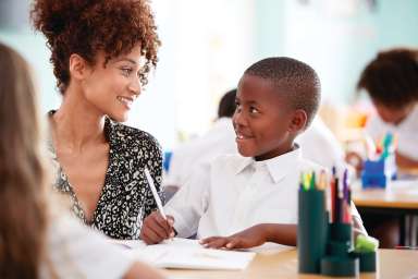 Woman Elementary School Teacher Giving Male Pupil Wearing Uniform One To One Support In Classroom