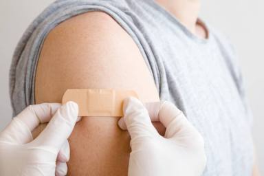 Doctor in white rubber protective gloves putting adhesive bandage on young man’s arm after scratch on skin or injection of vaccine. First aid. Medical, pharmacy and healthcare concept. Closeup.