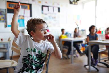 Cute boy with raised hand in classroom