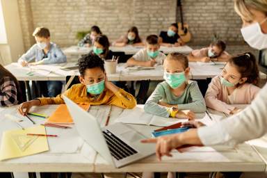 Elementary students wearing face masks while using laptop with their teacher in the classroom.