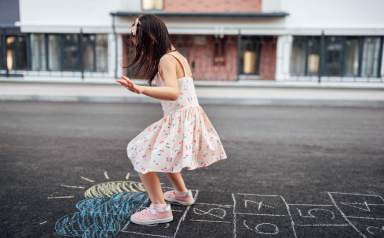 Image of cute little girl playing hopscotch on playground outdoors. Pretty child plays next to the house oustside. Kid plays hopscotch drawn on pavement. Summer activities and games for children.