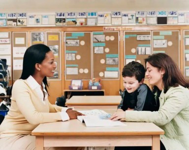 Teacher Sitting at a School Desk Showing a Book to a Parent and Her Son